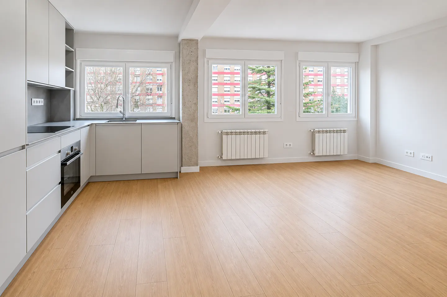 Modern kitchen with light grey cabinets, wood-look flooring, and large windows overlooking a residential building.