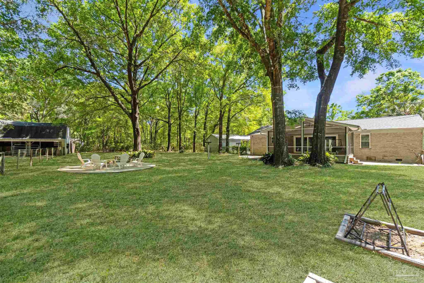 A large green lawn with trees, a patio with white chairs, and a brick house in the background.