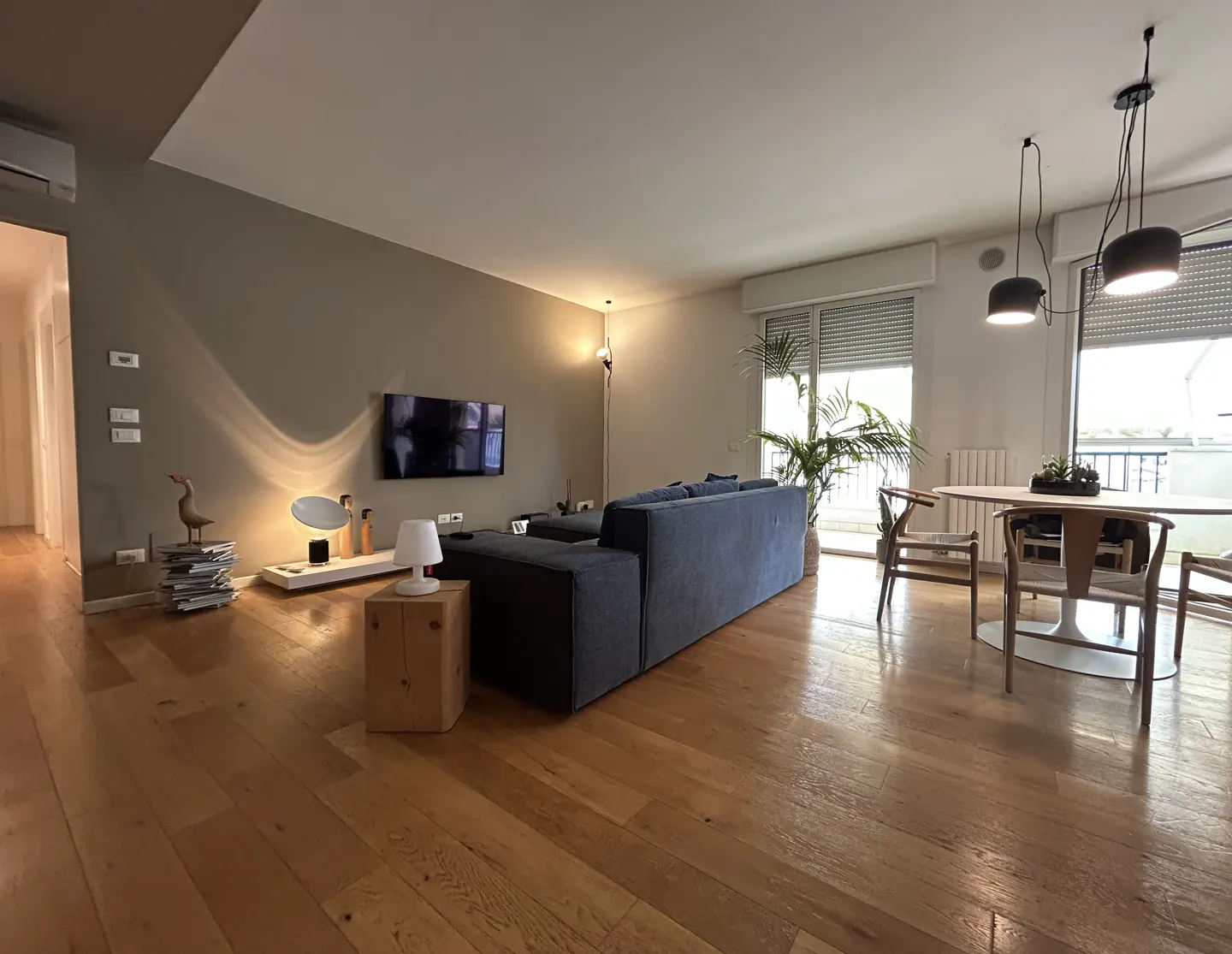 A modern living room with hardwood floors, a blue sectional sofa, a round dining table with chairs, and a TV on the wall.