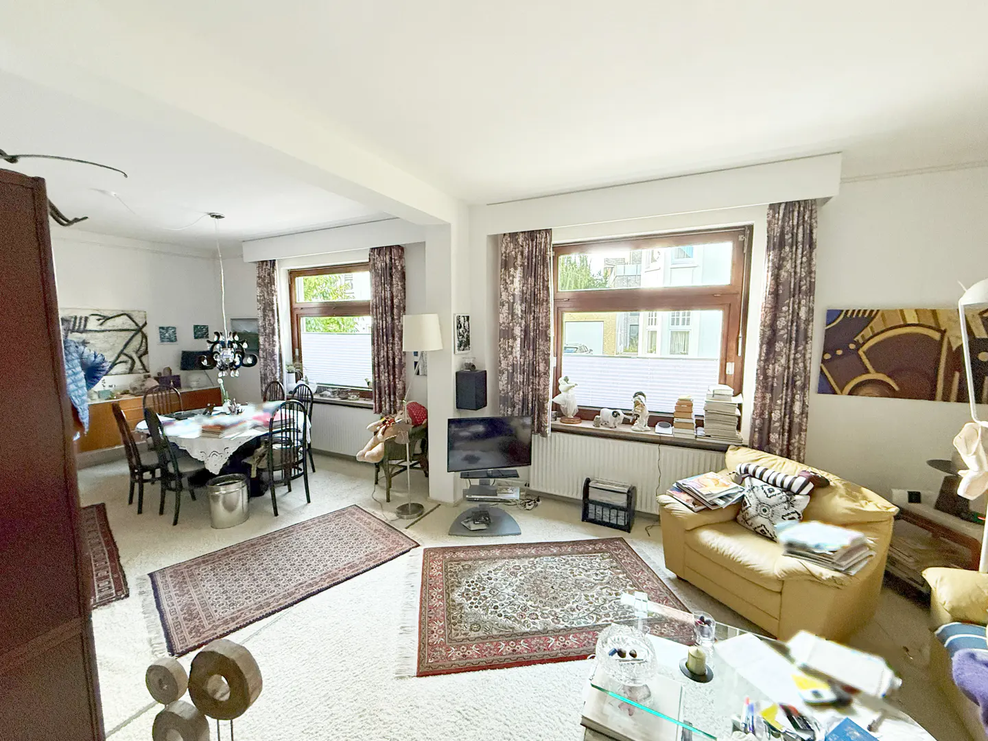 Bright living and dining room with white carpet, floral curtains, and large windows. A dining table and yellow armchair furnish the space.