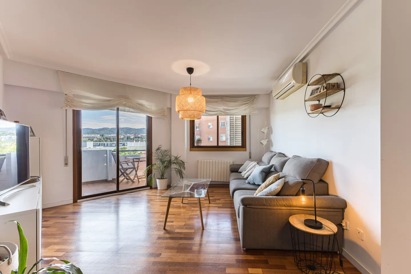 Bright living room with wood floors, a gray sofa with pillows, and a balcony with a view. A woven pendant light hangs above.