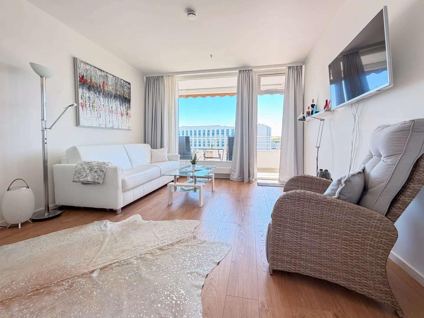 Bright living room with white sofa, wicker armchair, glass coffee table, abstract wall art, and balcony access.