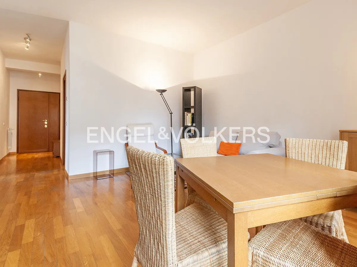 Bright living room with wood floors, a light wood table and woven chairs. A gray sofa with an orange pillow is in the background.