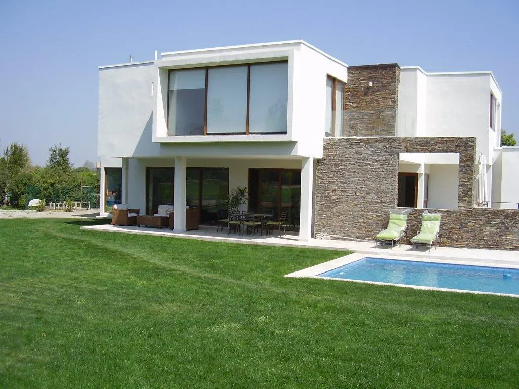 Two-story modern white house with stone accents, a pool, and a green lawn on a sunny day.