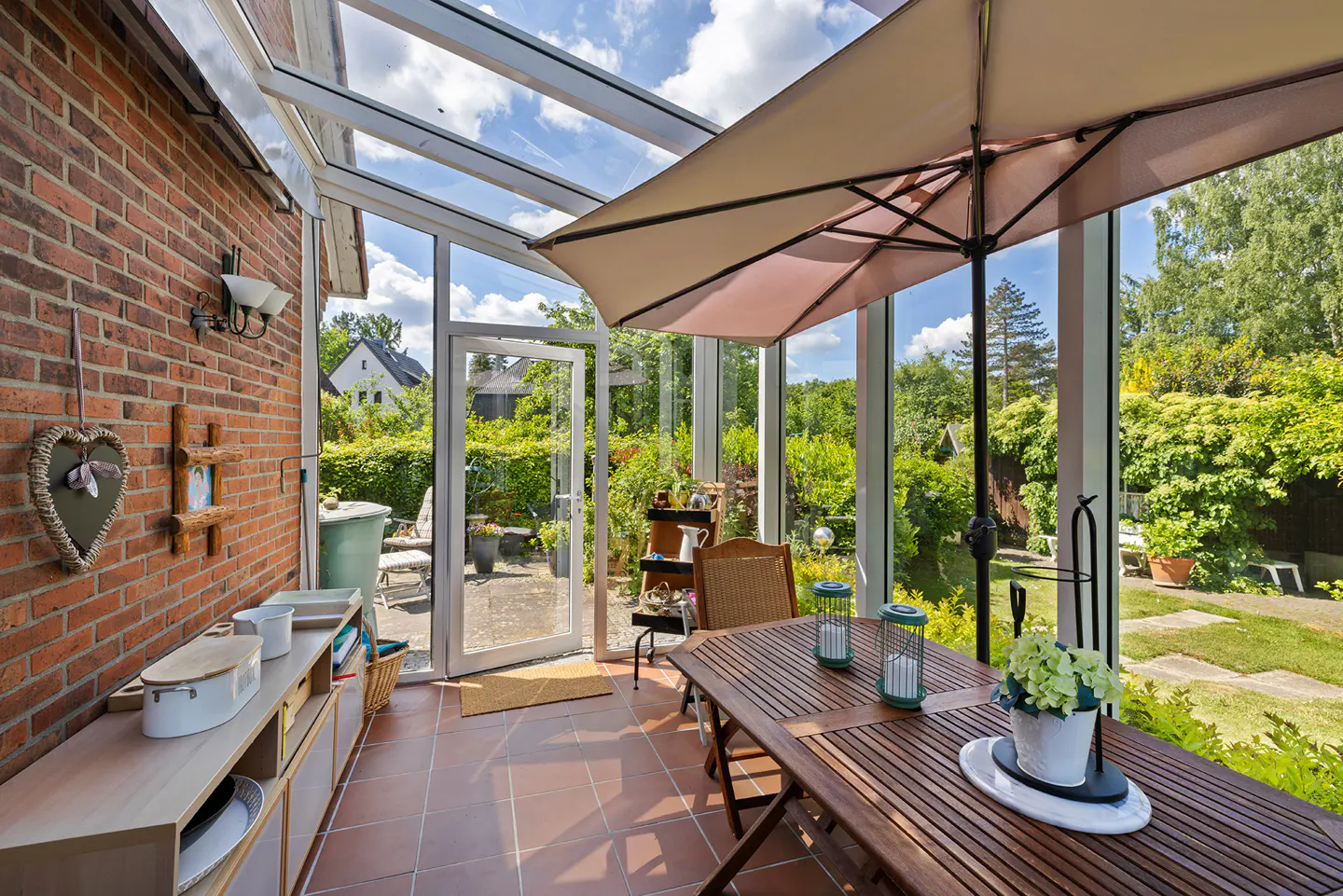 Sunroom with brick wall, glass ceiling, and open door to a lush green garden. A wooden table and umbrella provide shade.