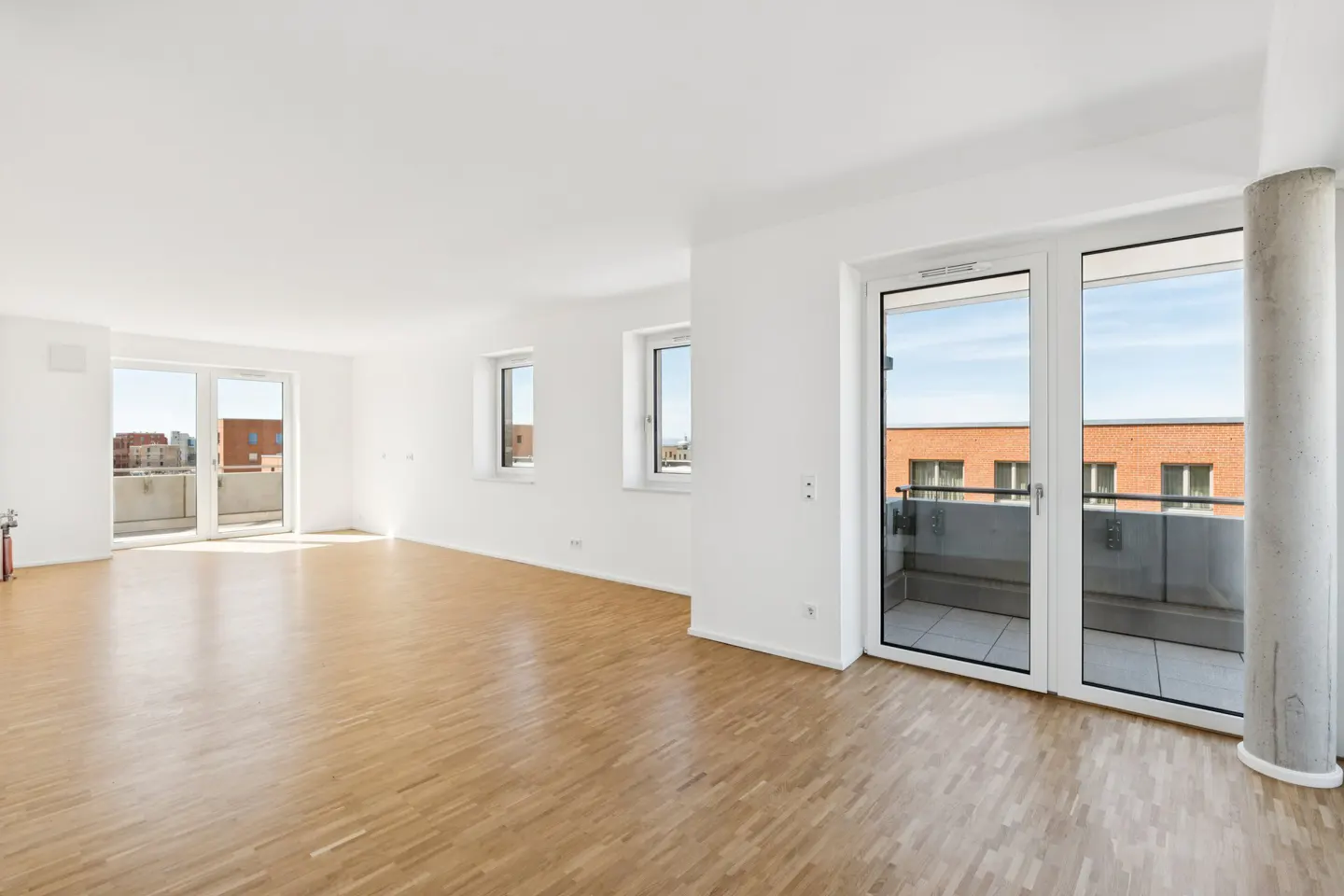 Bright, empty room with light wood floors, white walls, and large windows leading to balconies. A concrete pillar stands near one window.