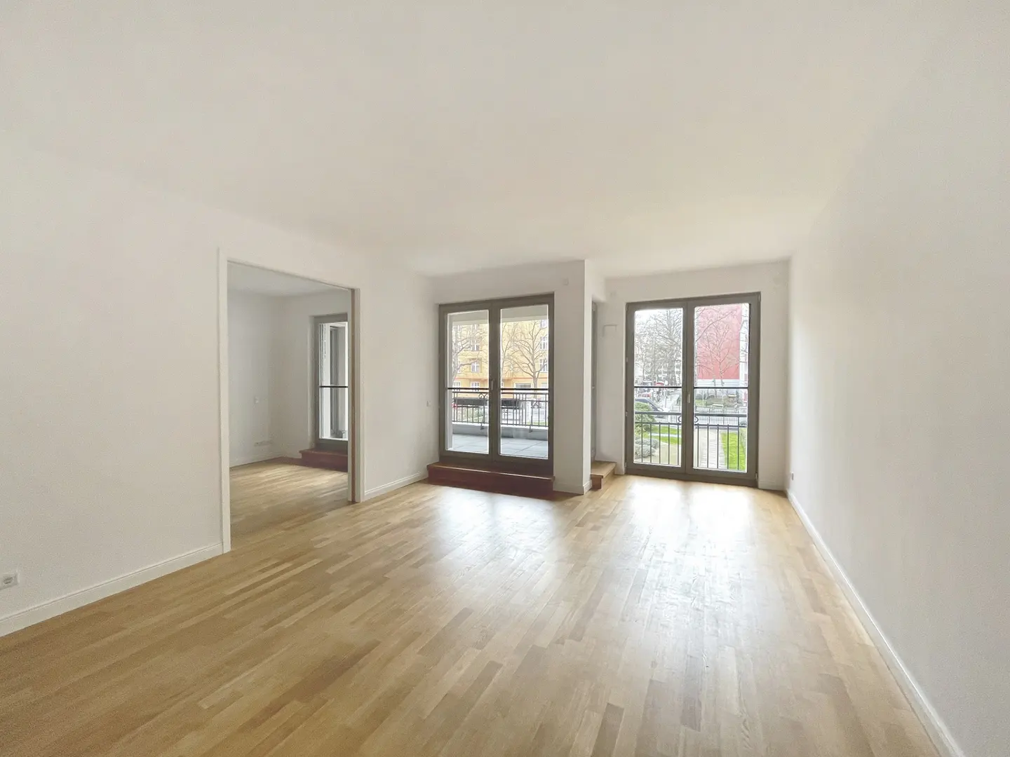 Bright, empty room with light wood floors and white walls. Two sets of glass doors lead to balconies. An open doorway leads to another room.