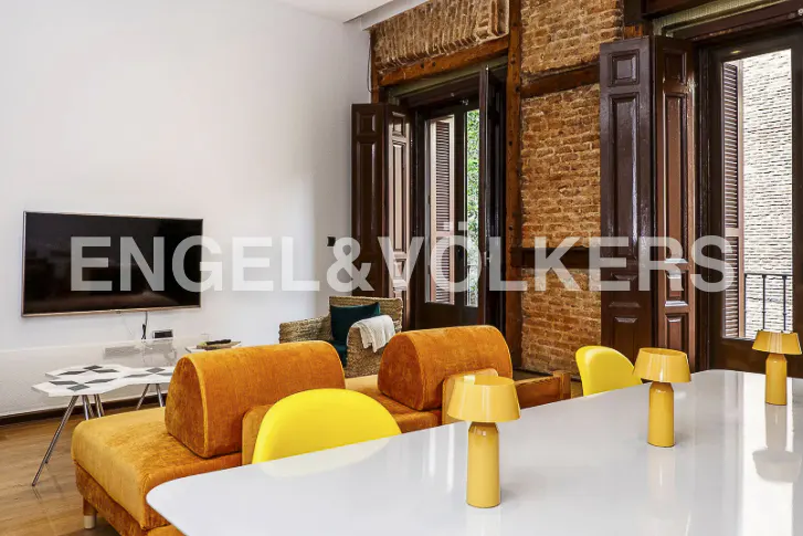 Living room with orange sofa, yellow chairs, and white table. Brick wall and dark wood window frames in the background.