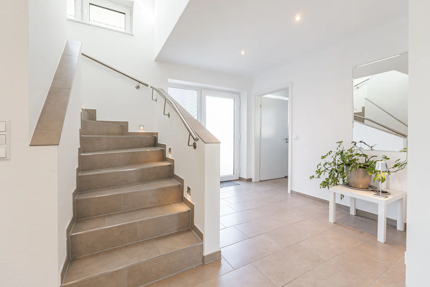 Bright foyer with tile floors, white walls, and a staircase with a metal handrail. A plant sits on a white table near a mirror.