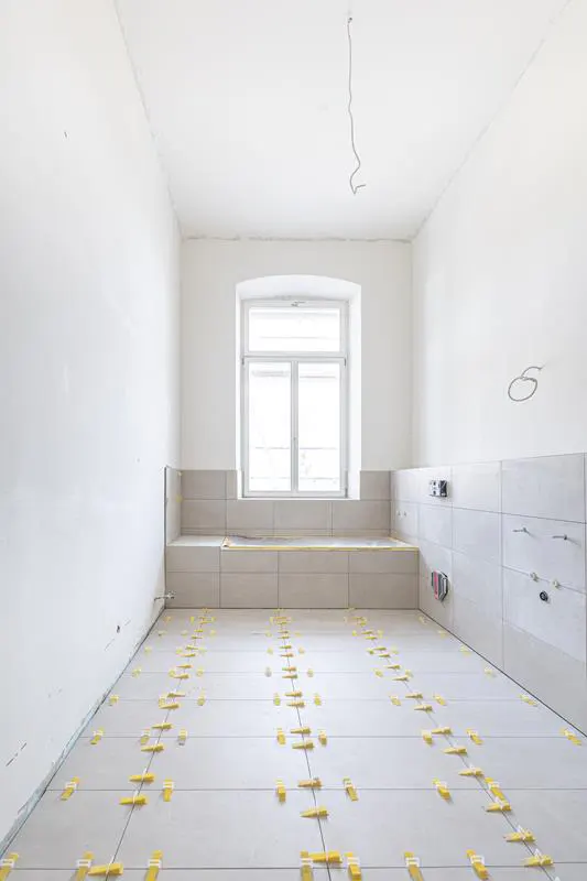 Bright, empty room with white walls and tiled floor. Yellow spacers are between the tiles. A window is centered on the back wall.