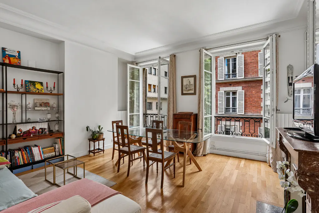 Bright living room with a glass dining table, wooden chairs, a bookshelf, and open windows overlooking a brick building.