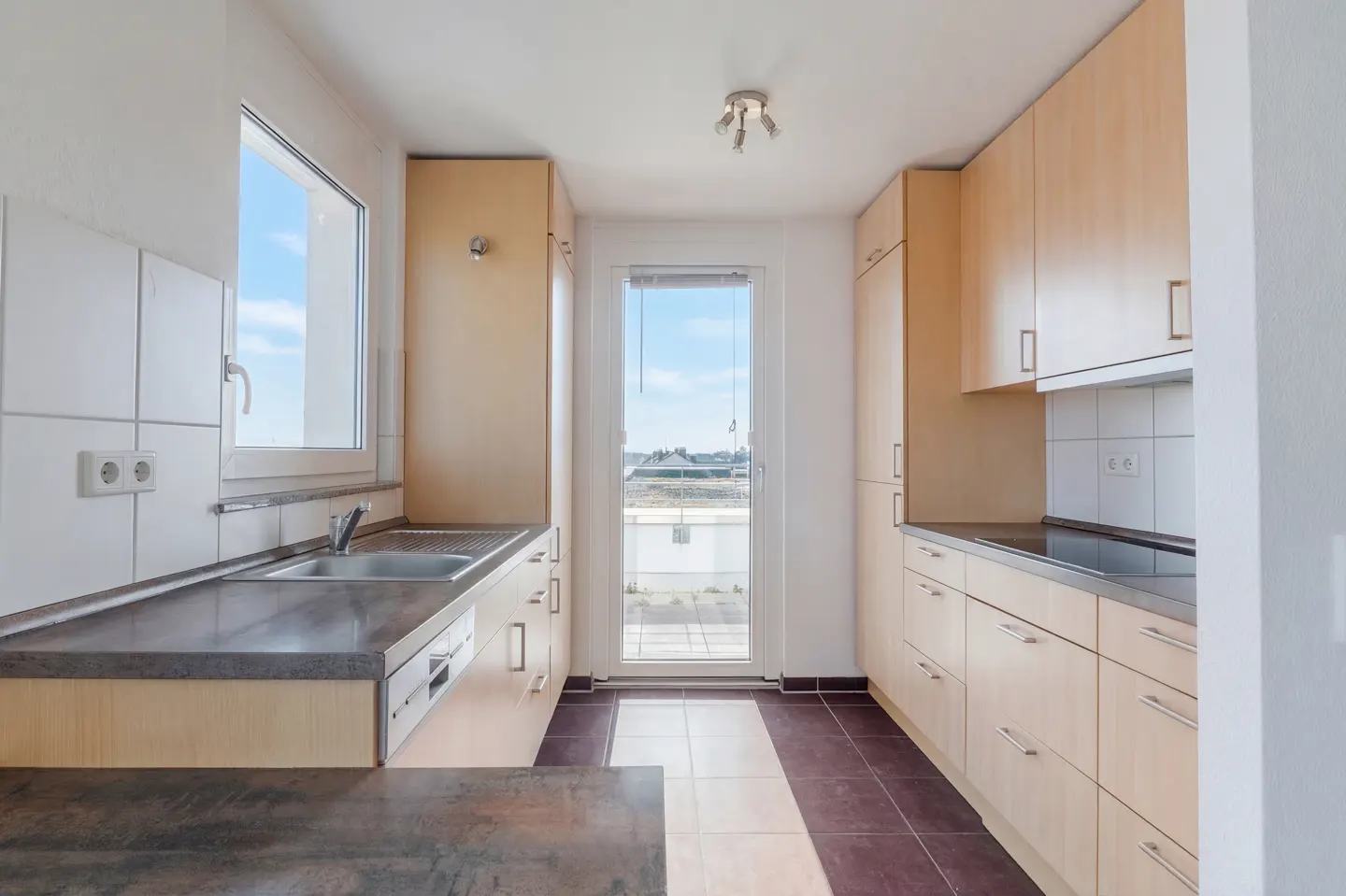 A galley kitchen with light wood cabinets, dark countertops, and a stainless steel sink under a window with a view of a blue sky.