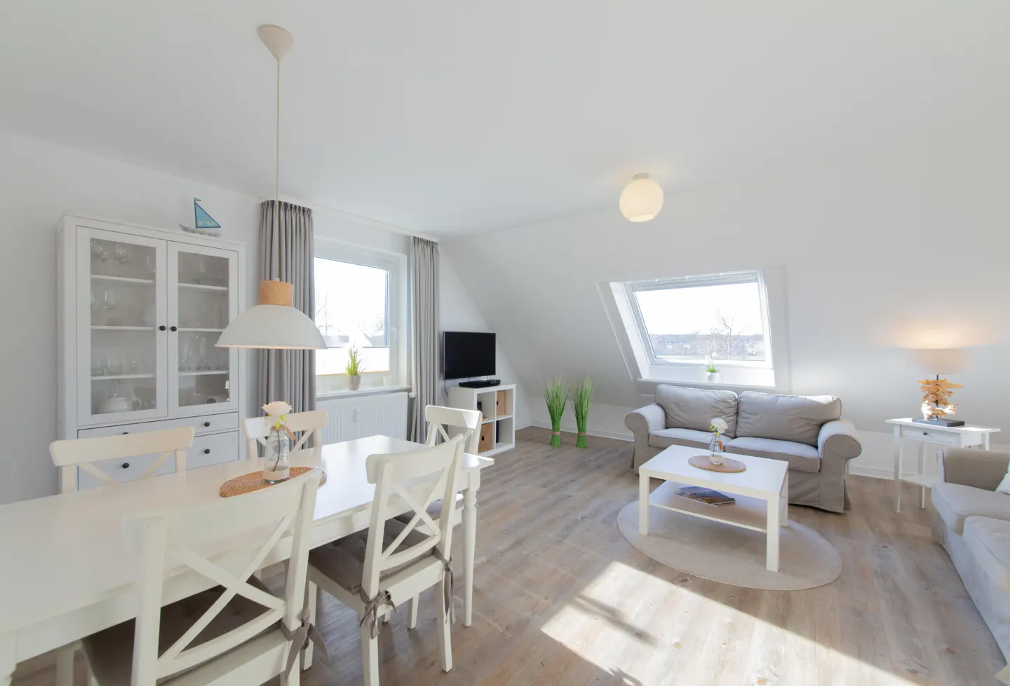 Bright, airy living space with white dining set, gray sofas, and wood floors under a sloped ceiling with skylight.