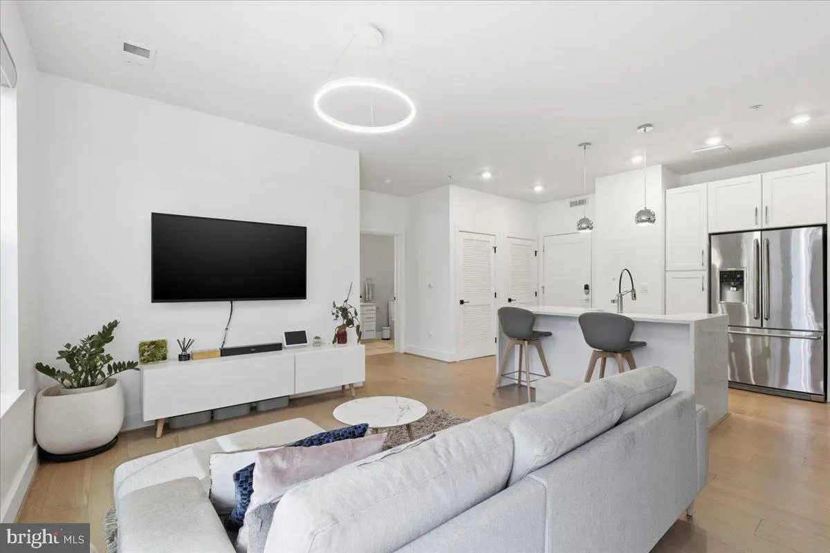 Bright, open-concept living space with white walls, light wood floors, and modern furnishings. A large TV is mounted above a white console table.
