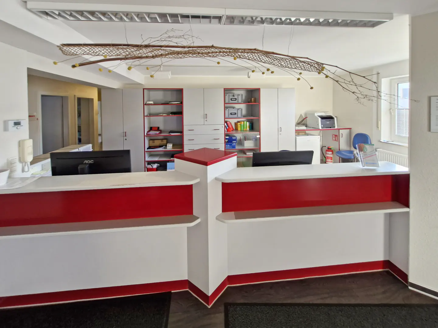 Modern office reception desk with red and white accents, featuring storage cabinets, computers, and a decorative branch.