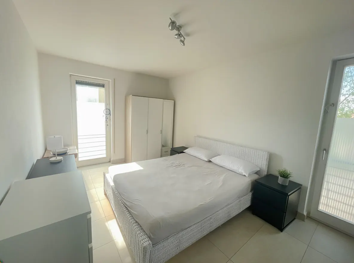 Bright bedroom with a white bed, two pillows, and a black nightstand with a plant. A white wardrobe and a door to the outside are visible.