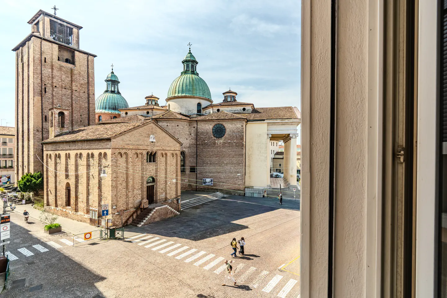 View from a window overlooking a historic Italian square with a brick church, a tall bell tower, and green-domed roofs.