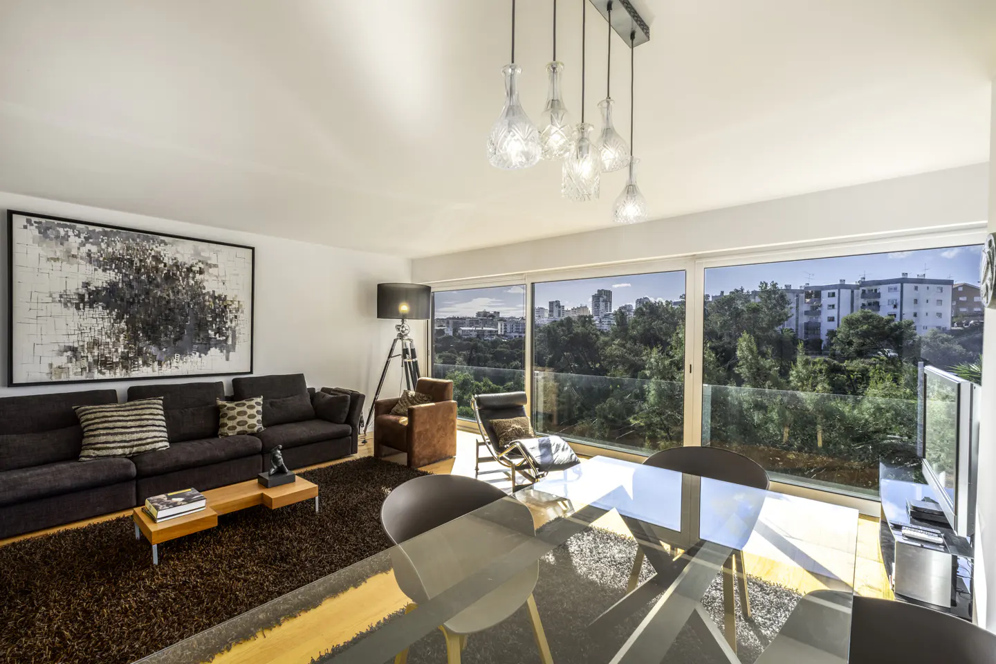 Bright living room with a brown sofa, glass table, and city view through large windows. Modern chandelier hangs above.