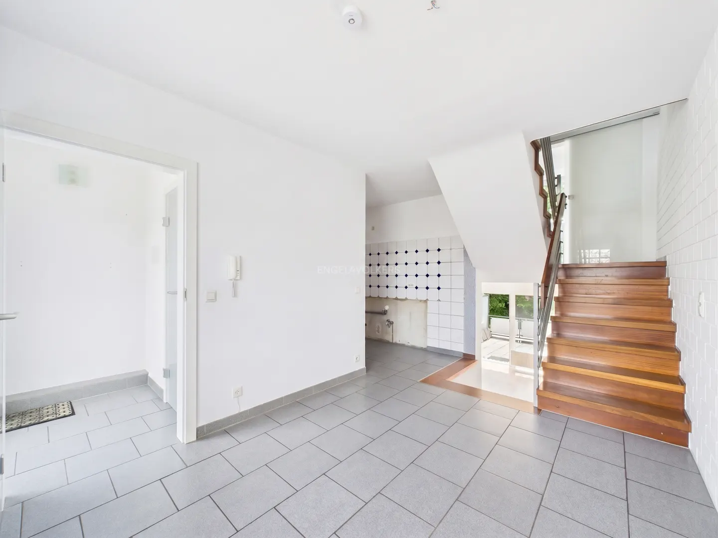 Bright, empty room with gray tile floor, white walls, and wooden stairs. Kitchen area visible in the background.