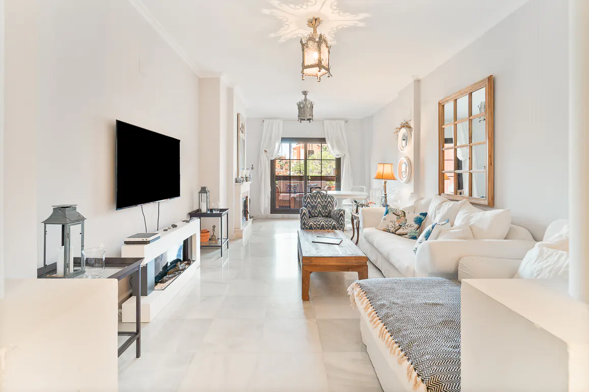 Bright living room with white walls, marble floors, and a large window. A TV is mounted on the wall, and a white sofa sits in the center.