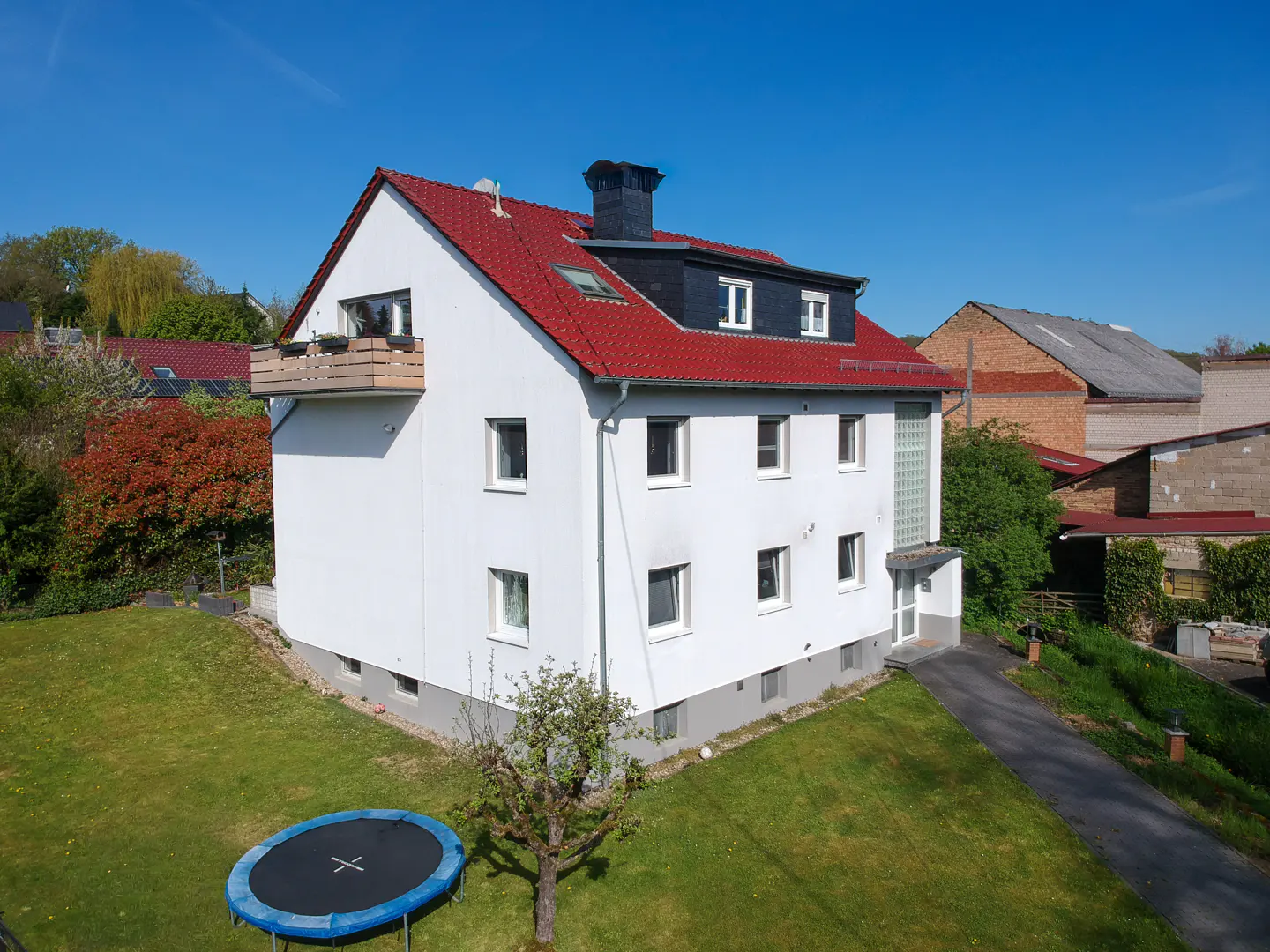 A three-story white house with a red roof and a black chimney stands on a green lawn under a blue sky. A trampoline is in the yard.