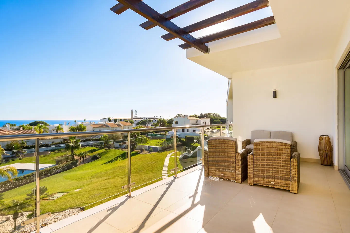 Balcony with wicker furniture overlooks a green lawn, houses, and the ocean under a blue sky.