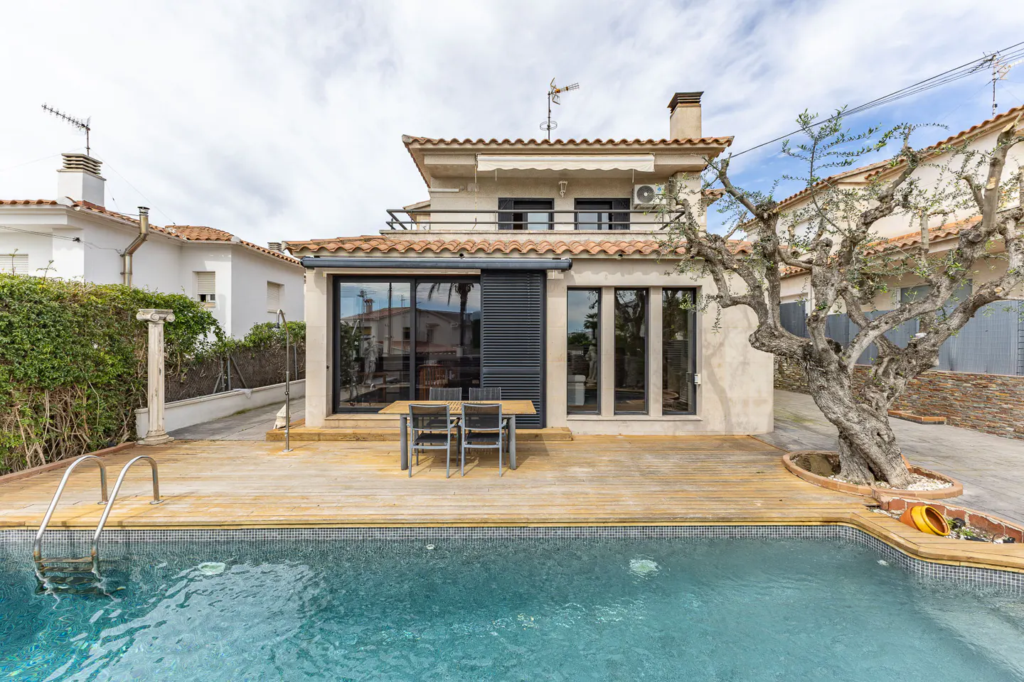 Backyard view of a two-story house with a pool, wooden deck, table, chairs, and a large tree.