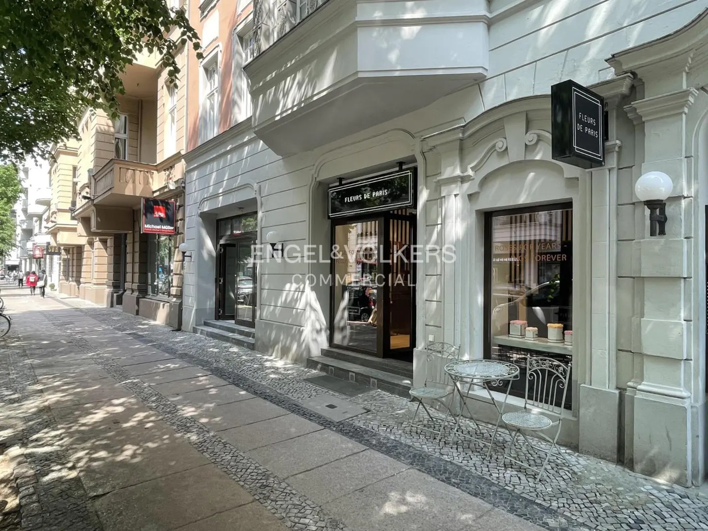 Exterior view of a light gray building with "Fleurs de Paris" signage and Engel & Volkers Commercial logo. A small table and chairs sit outside.