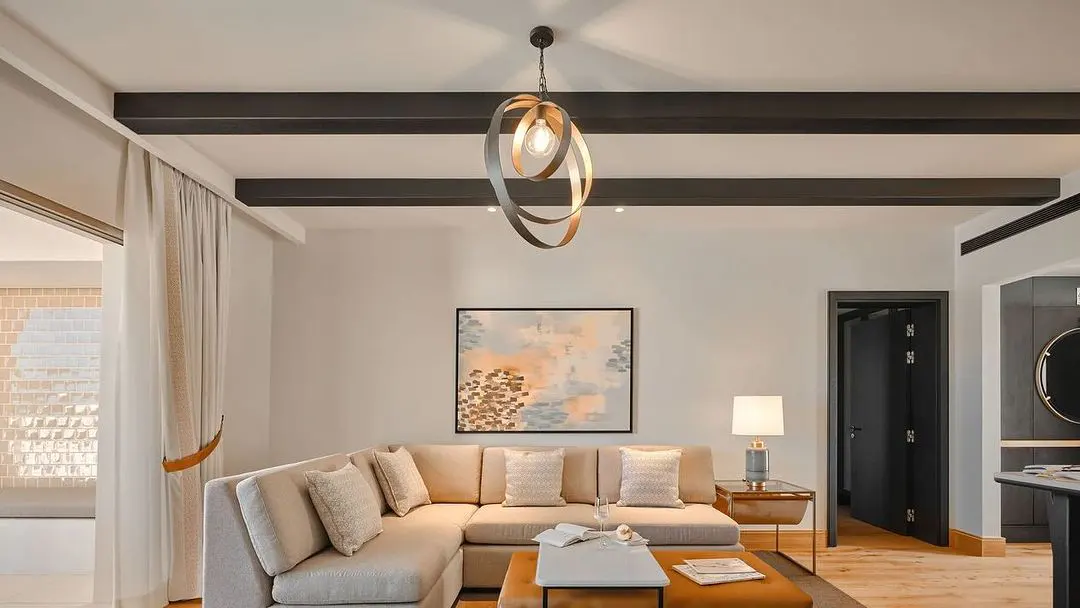 Living room with beige sectional sofa, abstract art, and modern light fixture. Black beams accent the ceiling.