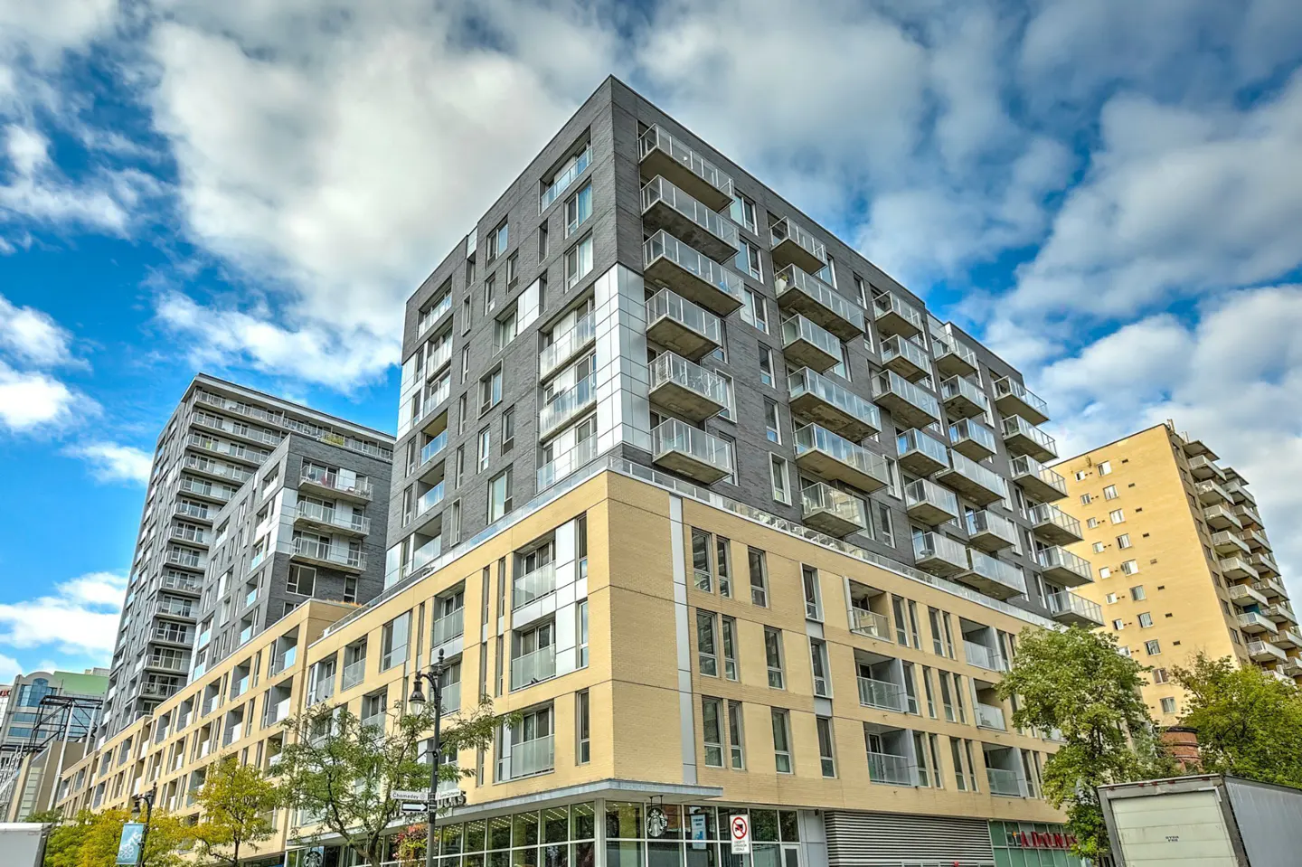Modern apartment buildings with grey and tan facades, glass balconies, and street-level retail under a blue, cloudy sky.