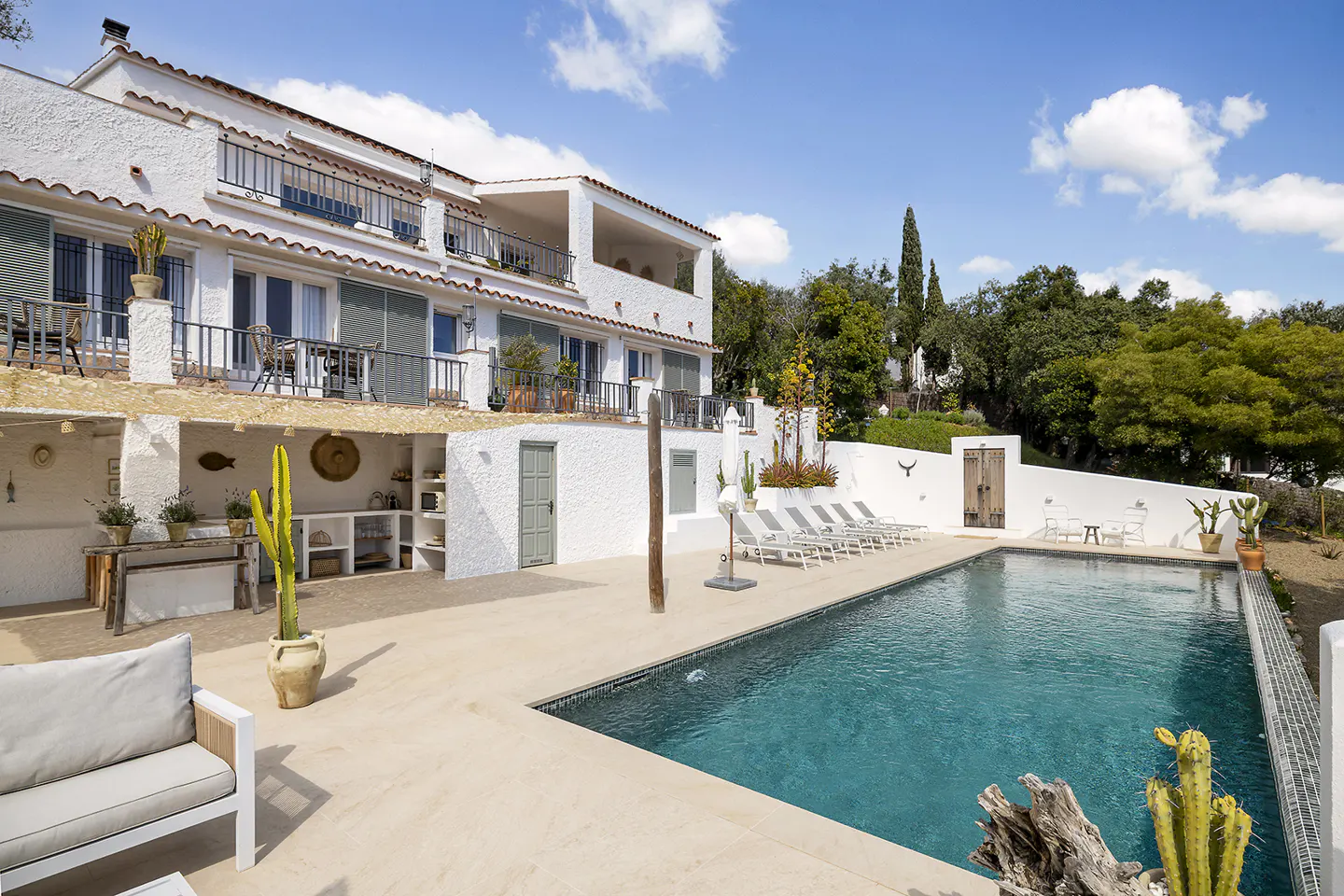 Exterior view of a white stucco house with a pool, lounge chairs, and cacti under a blue sky.