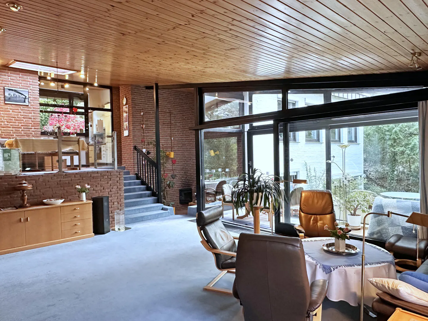 A spacious living room with wood ceilings, brick walls, and large windows. Leather chairs and a table with flowers are in the foreground.