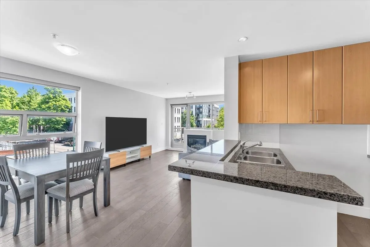 Bright, open-concept living space with gray wood floors. Kitchen has granite counters and wood cabinets. Dining table and TV in the living area.