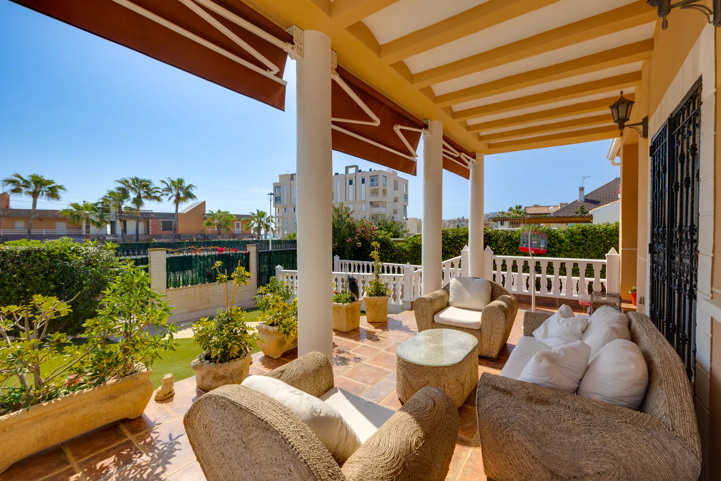 Covered patio with wicker furniture, white cushions, and potted plants. View of a green lawn, palm trees, and blue sky.