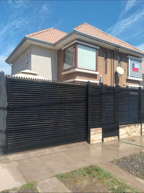 Two-story house with a brown roof and beige walls behind a black horizontal slat fence. A Chilean flag is visible on the right.