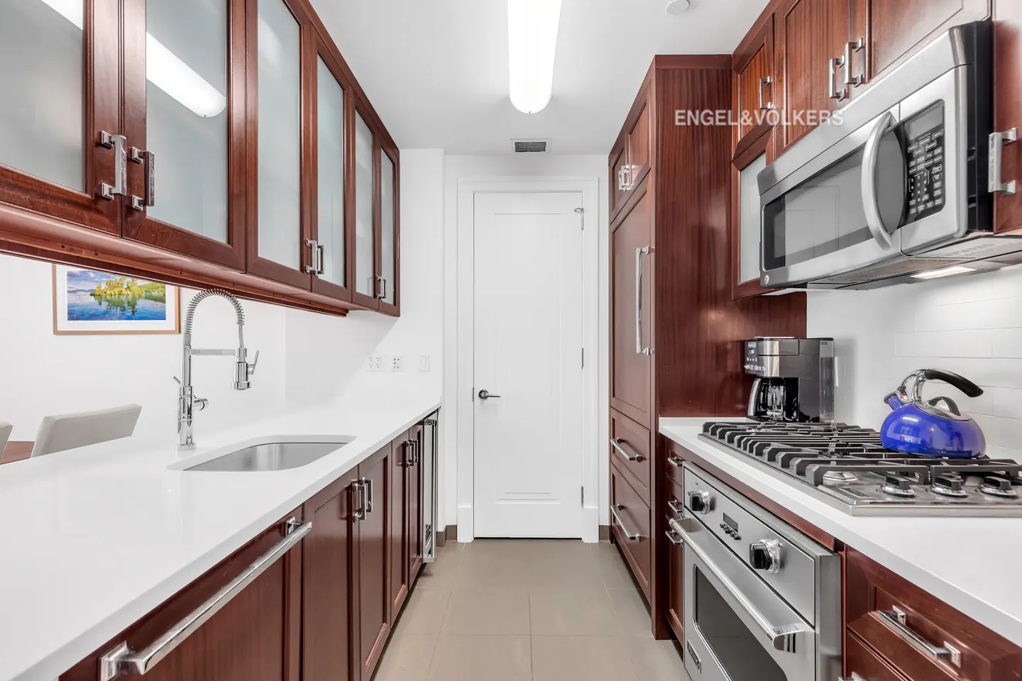 A galley kitchen with dark wood cabinets, white countertops, and stainless steel appliances. A white door is at the end of the kitchen.