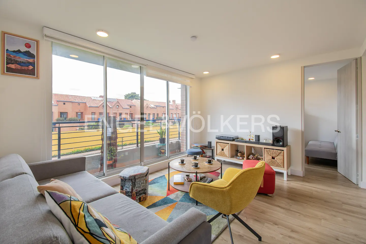 Modern living room with a gray sofa, yellow armchair, colorful geometric rug, and large windows overlooking a terrace.