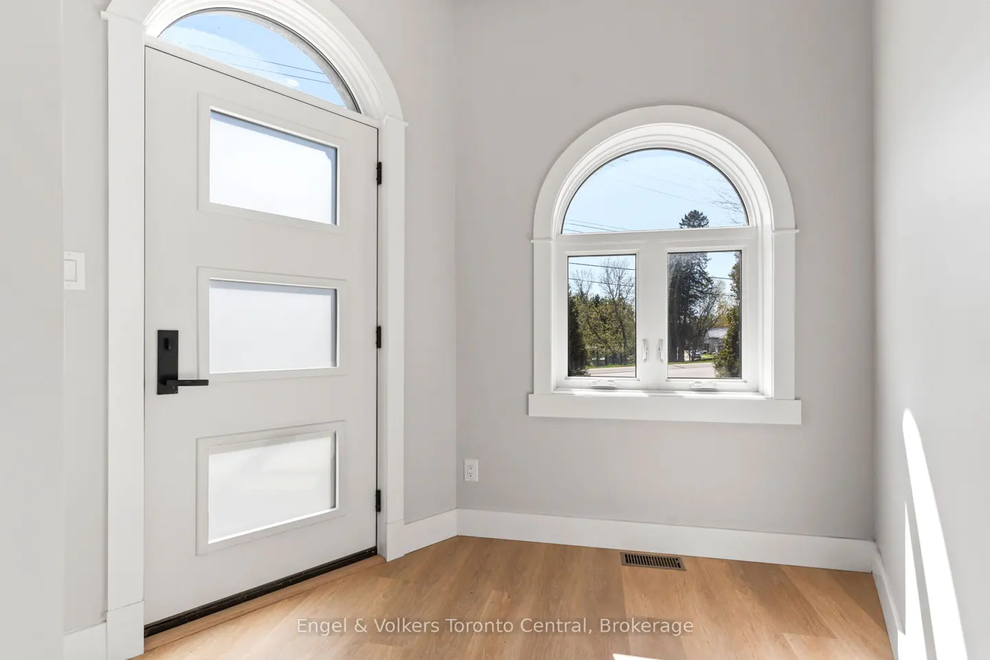 Entryway with a white door with three glass panels, a black handle, and an arched window. The walls are light gray, and the floor is light wood.