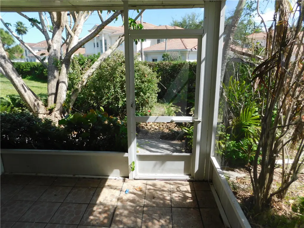 View from a screened-in porch with a white door open to a lush green garden with trees and bushes.