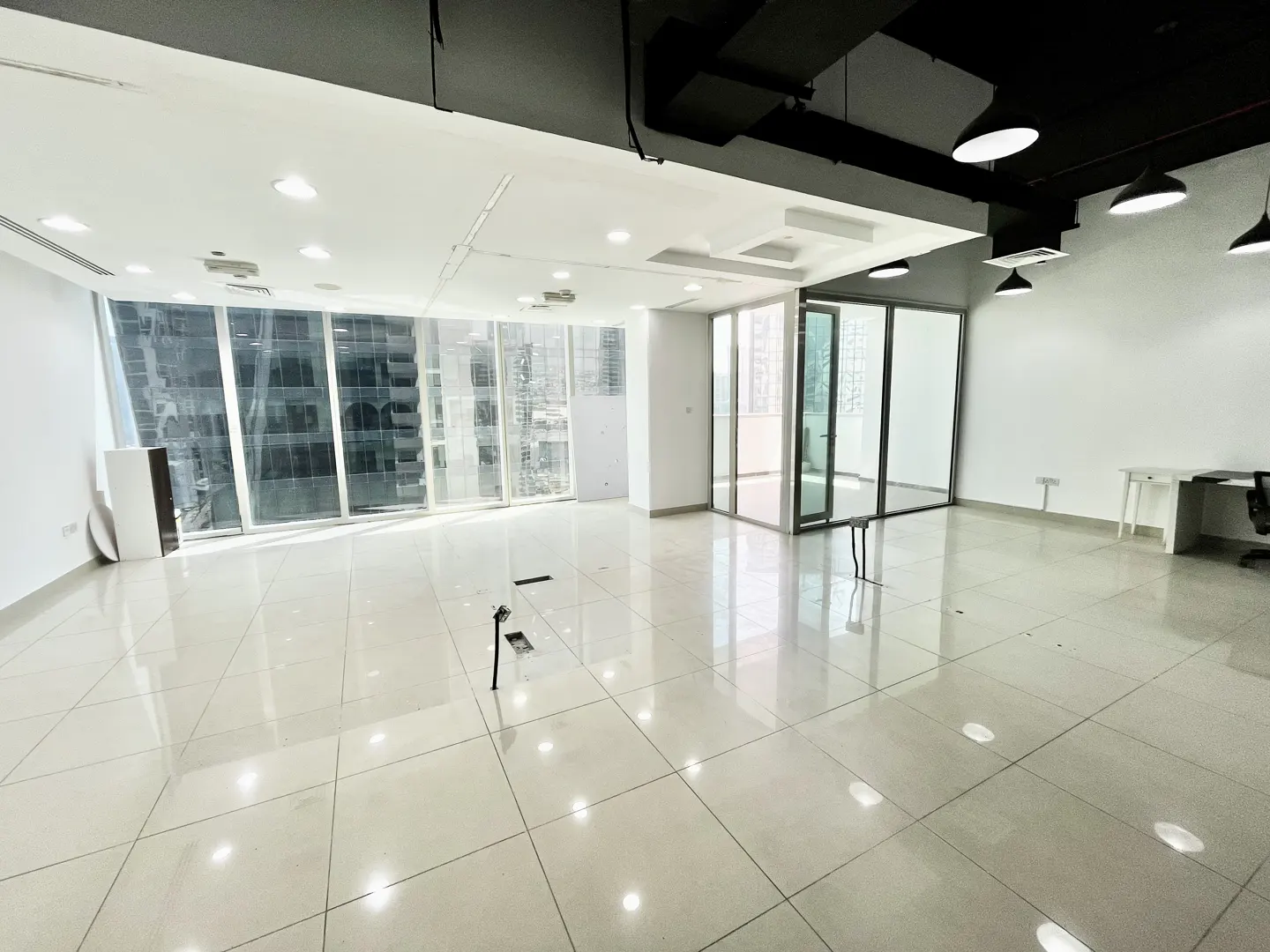 Bright, empty office space with white walls, tile floor, and large windows showing city views. Black ceiling fixtures and a glass-walled room are visible.
