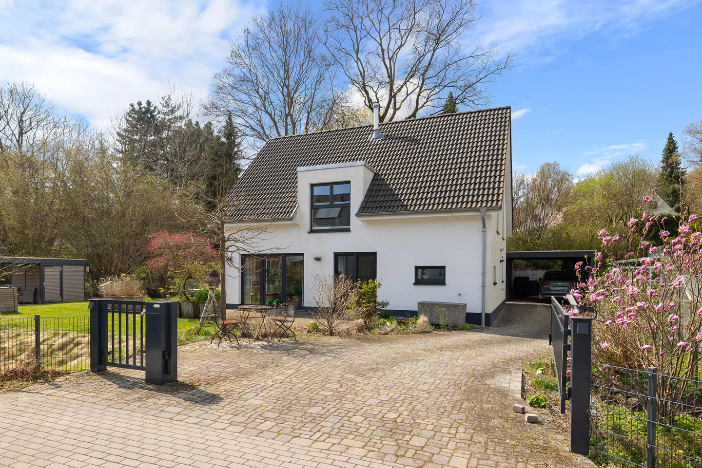 Exterior view of a two-story white house with a dark gray roof and black window frames. A paved driveway leads to a carport.