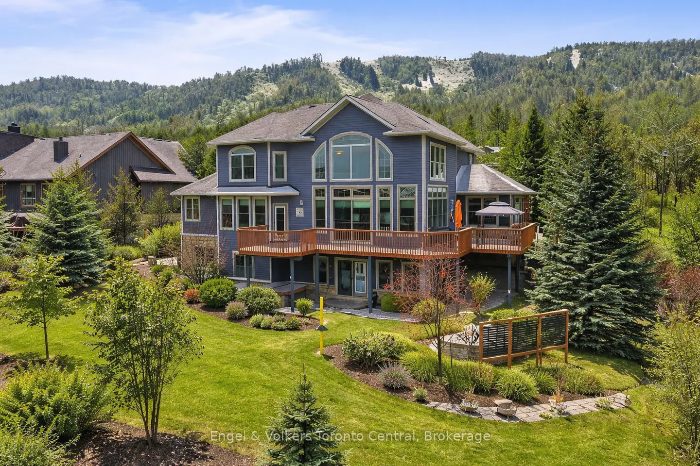 Blue two-story house with a brown deck, surrounded by green trees and grass. A mountain is in the background.