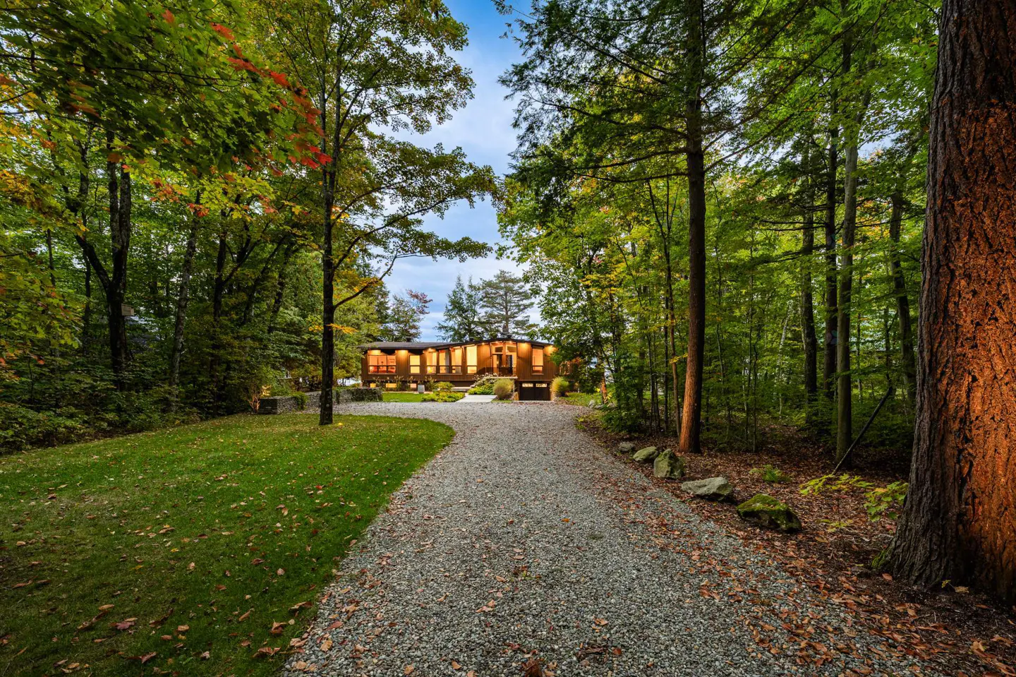 A gravel driveway leads to a modern brown house surrounded by green trees and grass.