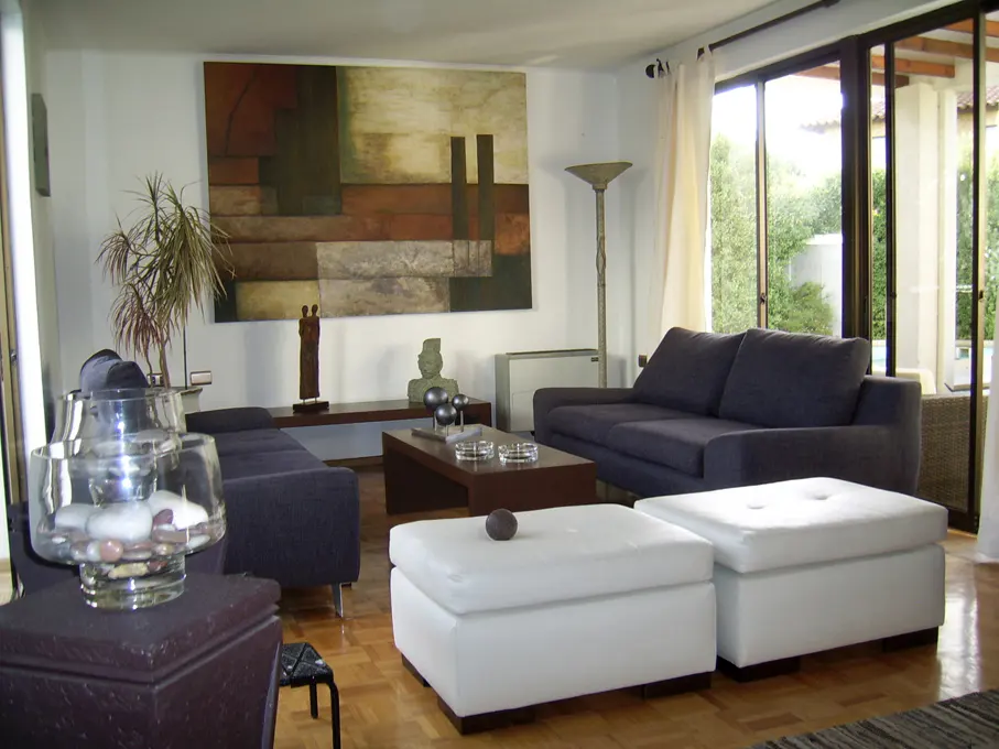 A living room with blue sofas, white ottomans, and a large abstract painting on the wall. A sliding glass door leads to a patio.