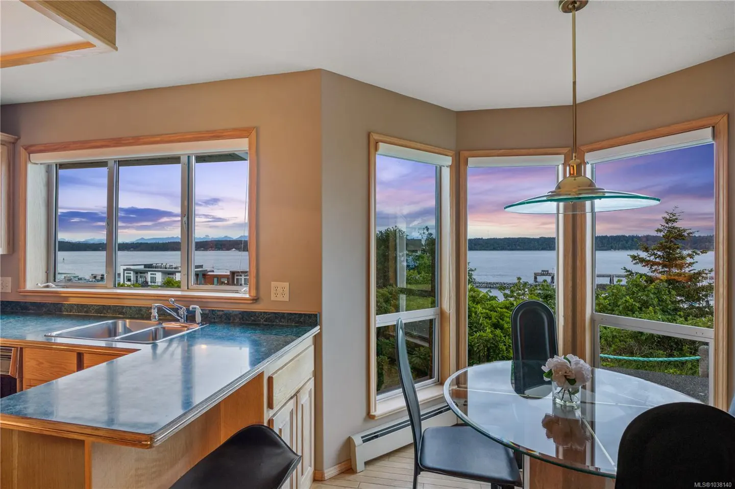 A kitchen and dining area with a view of the water at sunset. A glass table with black chairs sits under a pendant light.