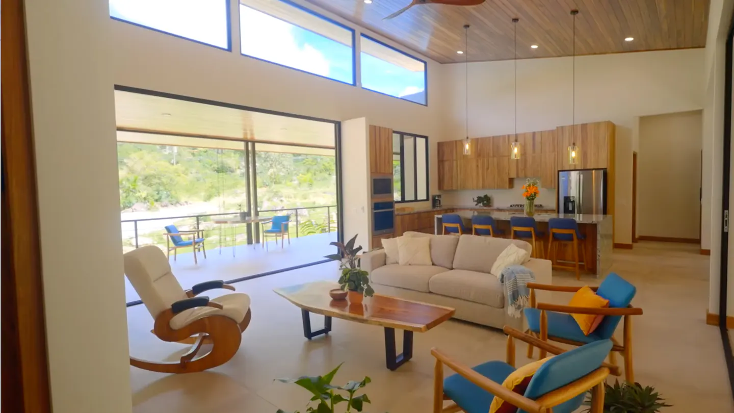 A bright, open-concept living space with a beige sofa, wooden chairs, and a kitchen with wooden cabinets. Large windows offer a view of a green landscape.