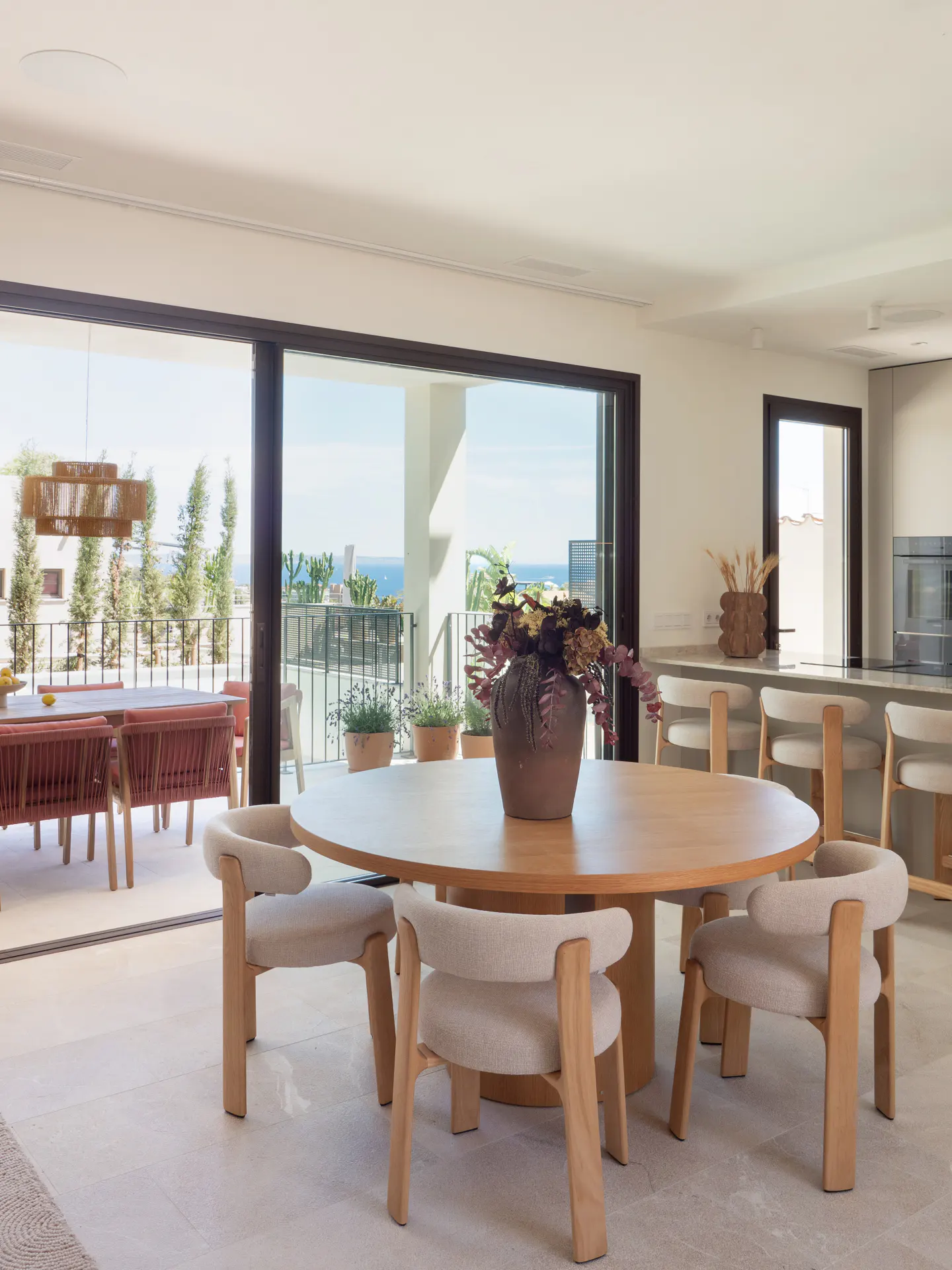 Bright, modern dining area with a round wooden table, four chairs, and a vase of flowers. Sliding glass doors open to a patio with ocean views.