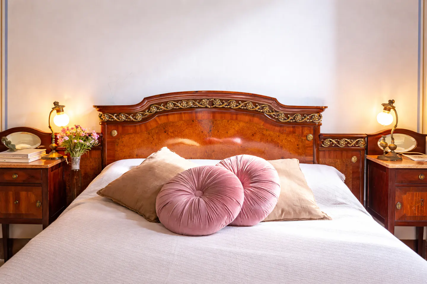 Bedroom with a wooden headboard, white bedding, and pink round pillows. Nightstands with lamps and flowers flank the bed.