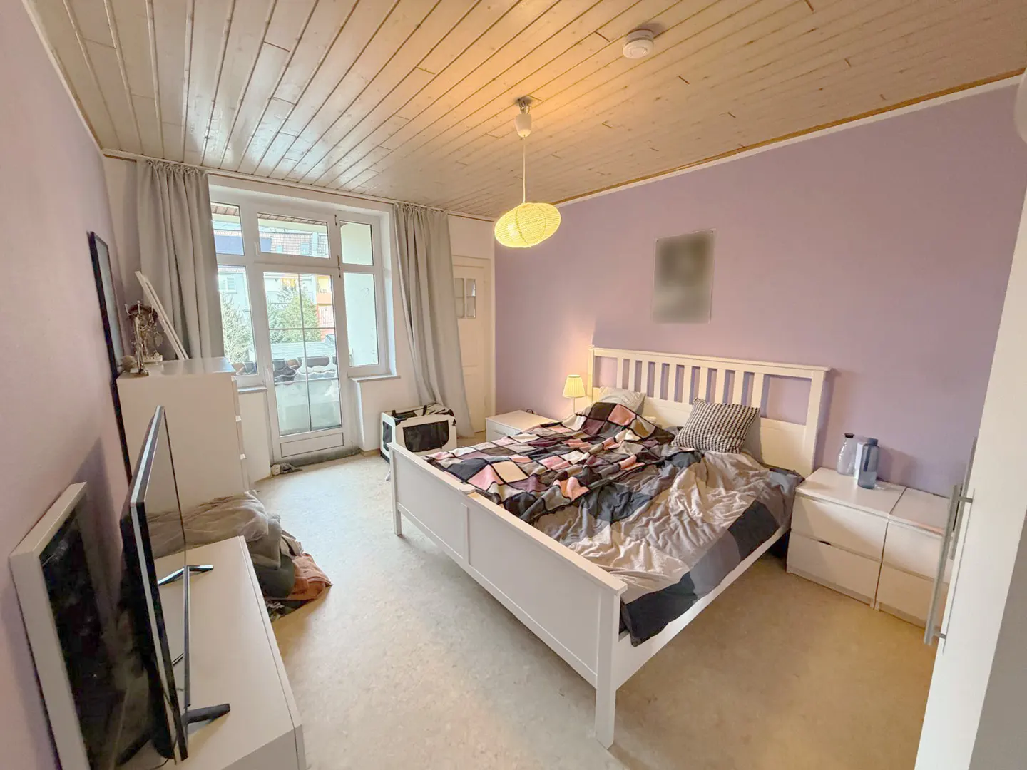 Bedroom with lilac walls, white wood ceiling, and a white bed with a patterned quilt. A TV and dresser are on the left.
