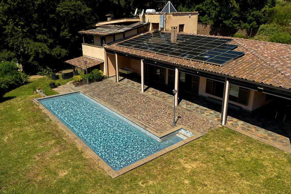 Aerial view of a house with a tiled roof, solar panels, a stone patio, and a rectangular swimming pool in a grassy yard.